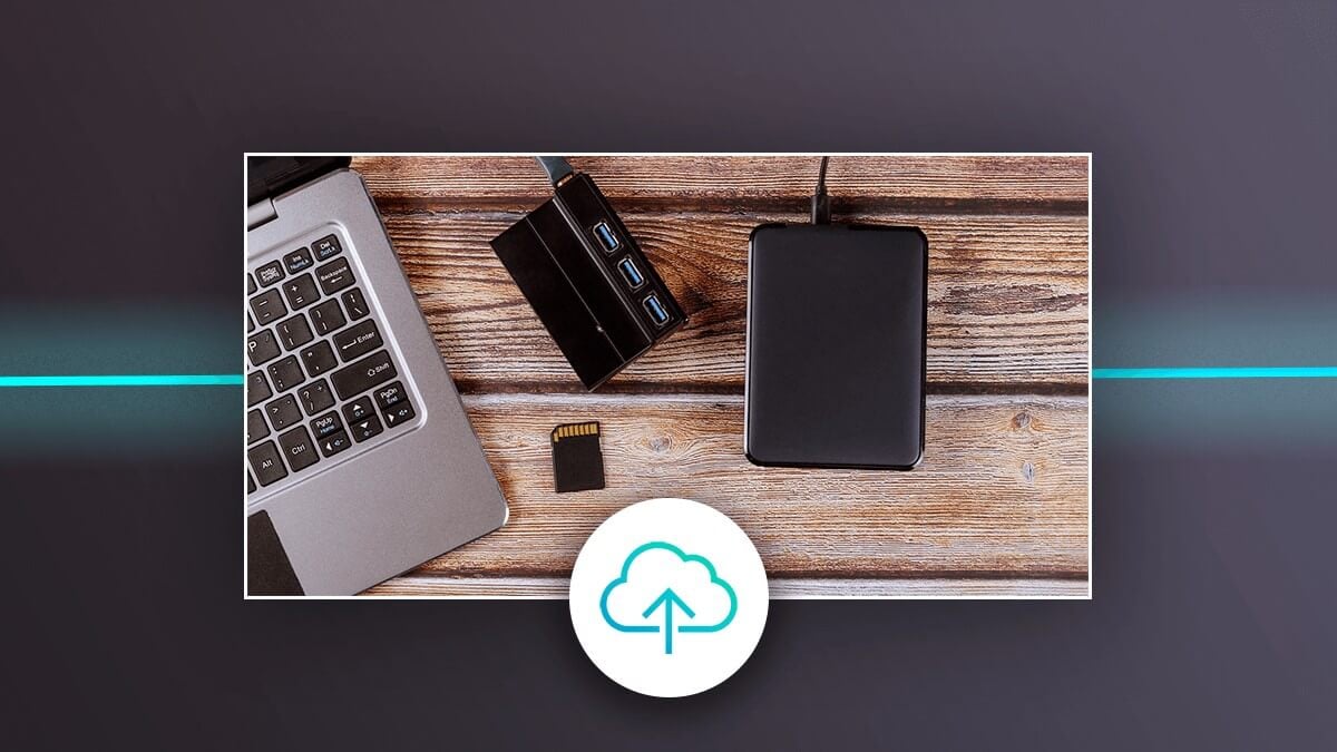 Laptop with external drive, USB hub, and memory card on a desk, with a cloud upload icon.
