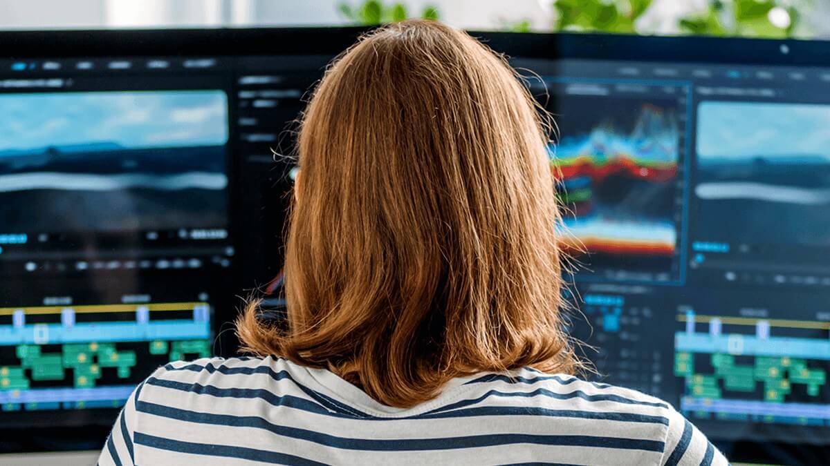 Person viewed from behind editing video on dual monitors with timeline and color correction tools visible.