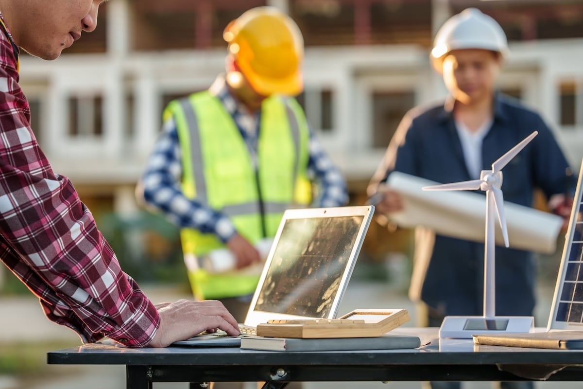 Engineer working on a laptop beside wind turbine and solar panel models while two construction workers review plans in the background.