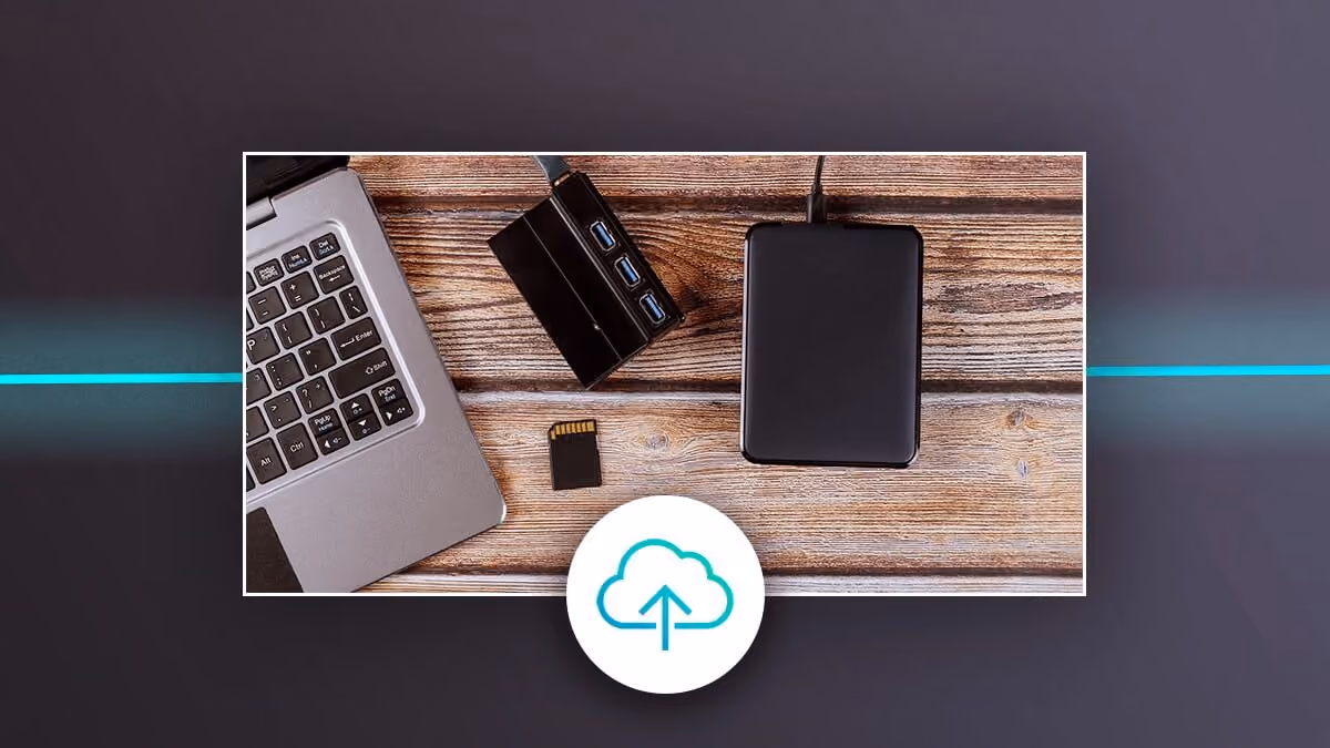 Laptop with external drive, USB hub, and memory card on a desk, with a cloud upload icon.